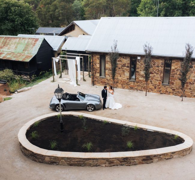 Bride and groom beside vintage convertible at rustic venue